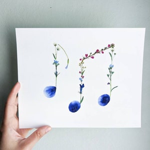 May include: A white card with a floral arrangement in the shape of musical notes. The notes are made of pressed flowers in shades of blue, pink, and white.
