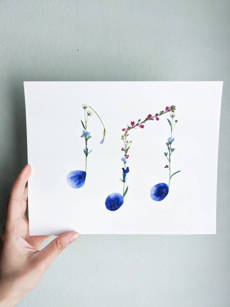 May include: A white card with a floral arrangement in the shape of musical notes. The notes are made of pressed flowers in shades of blue, pink, and white.