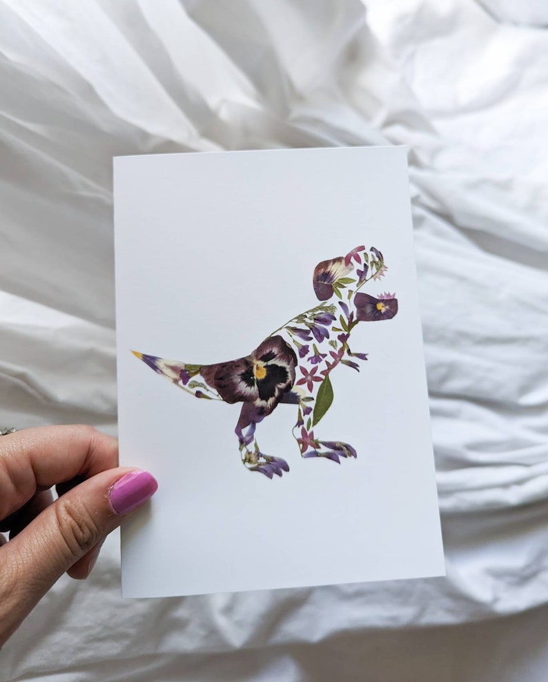 May include: A white card with a floral pressed flower design of a Tyrannosaurus Rex dinosaur. The flowers are purple, pink, and green.