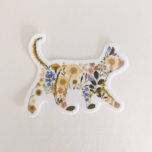 May include: A white sticker with the silhouette of a cat made of pressed flowers. The flowers are mostly yellow, white, and blue.