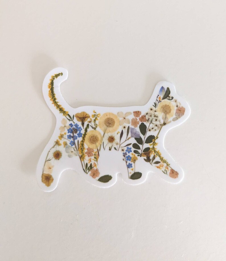 May include: A white sticker with the silhouette of a cat made of pressed flowers. The flowers are mostly yellow, white, and blue.