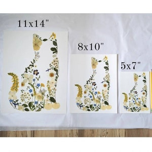 May include: Three different sizes of pressed flower art prints featuring a cat silhouette made of flowers. The prints are 11x14 inches, 8x10 inches, and 5x7 inches.