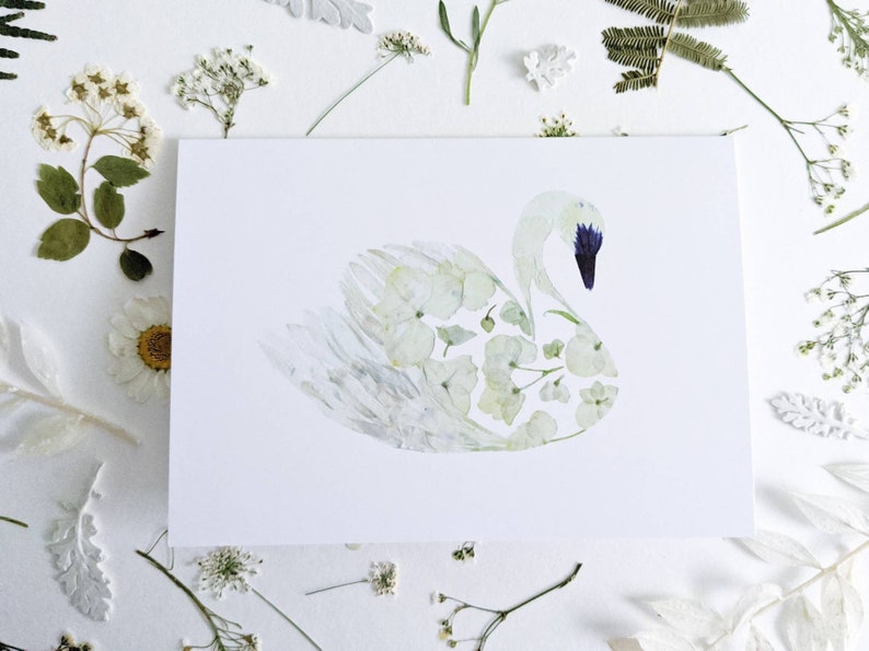 May include: A white greeting card featuring a swan illustration made of pressed flowers. The swan has a blue beak and is surrounded by dried flowers and leaves. The card is on a white surface.