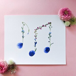 May include: A white card with three blue flower petals arranged in the shape of musical notes. The notes are made of pressed flowers and are surrounded by other small flowers in shades of pink, purple, and white.