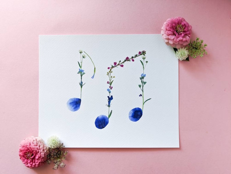 May include: A white card with three blue flower petals arranged in the shape of musical notes. The notes are made of pressed flowers and are surrounded by other small flowers in shades of pink, purple, and white.