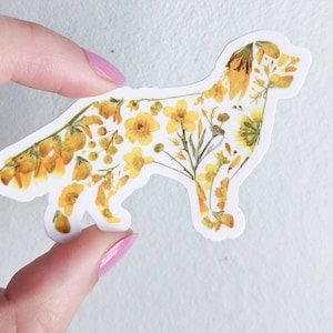 Puede incluir: Una pegatina blanca con la silueta de un perro delineada con flores amarillas prensadas.