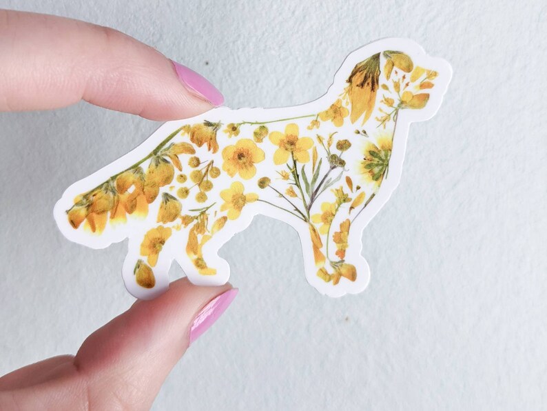 Puede incluir: Una pegatina blanca con la silueta de un perro delineada con flores amarillas prensadas.