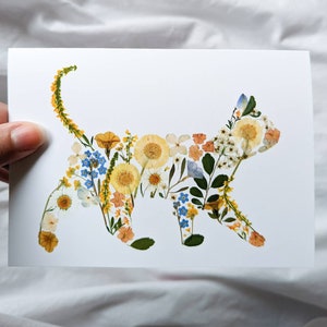 May include: A white card featuring a cat silhouette composed of dried flowers. The cat shape is filled with yellow, orange, blue, and white floral elements. The card is held by a hand against a white background.
