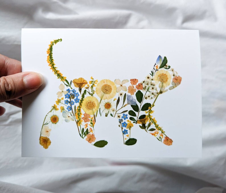 May include: A white card featuring a cat silhouette composed of dried flowers. The cat shape is filled with yellow, orange, blue, and white floral elements. The card is held by a hand against a white background.