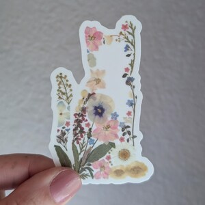 Puede incluir: Una pegatina blanca con un dise&ntilde;o floral con flores prensadas en tonos de rosa, azul y amarillo. Las flores est&aacute;n dispuestas en forma de gato.