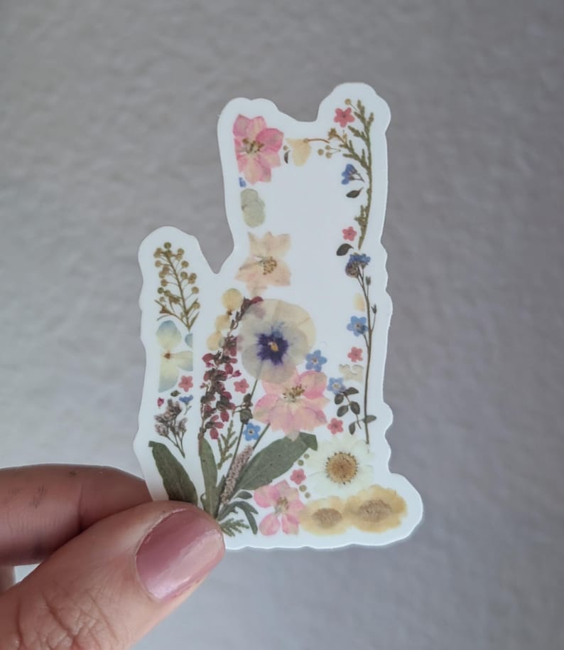 Puede incluir: Una pegatina blanca con un dise&ntilde;o floral con flores prensadas en tonos de rosa, azul y amarillo. Las flores est&aacute;n dispuestas en forma de gato.
