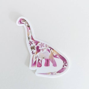 Op de afbeelding: Een witte sticker met een roze en wit bloemenontwerp in de vorm van een dinosaurus.