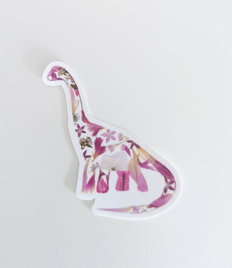 Op de afbeelding: Een witte sticker met een roze en wit bloemenontwerp in de vorm van een dinosaurus.
