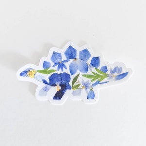 Op de afbeelding: Een blauwe en groene bloemen sticker van een stegosaurus dinosaurus. De dinosaurus is gemaakt van geperste bloemen en bladeren.