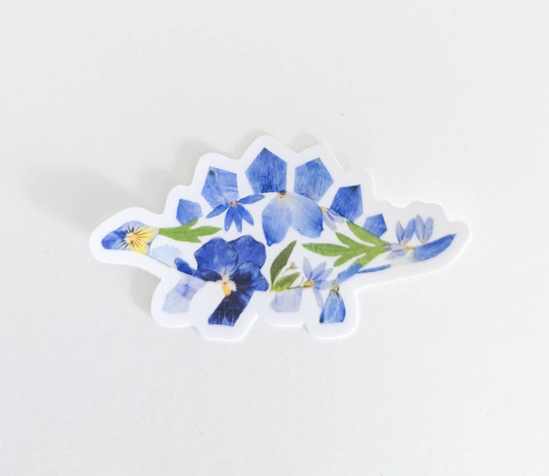 Op de afbeelding: Een blauwe en groene bloemen sticker van een stegosaurus dinosaurus. De dinosaurus is gemaakt van geperste bloemen en bladeren.