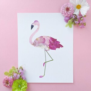 May include: A flamingo art print made from pressed pink and purple flower petals. The flamingo stands on one leg, with a black beak and a pink body. The artwork is on a white background, with pink flowers in the corners.