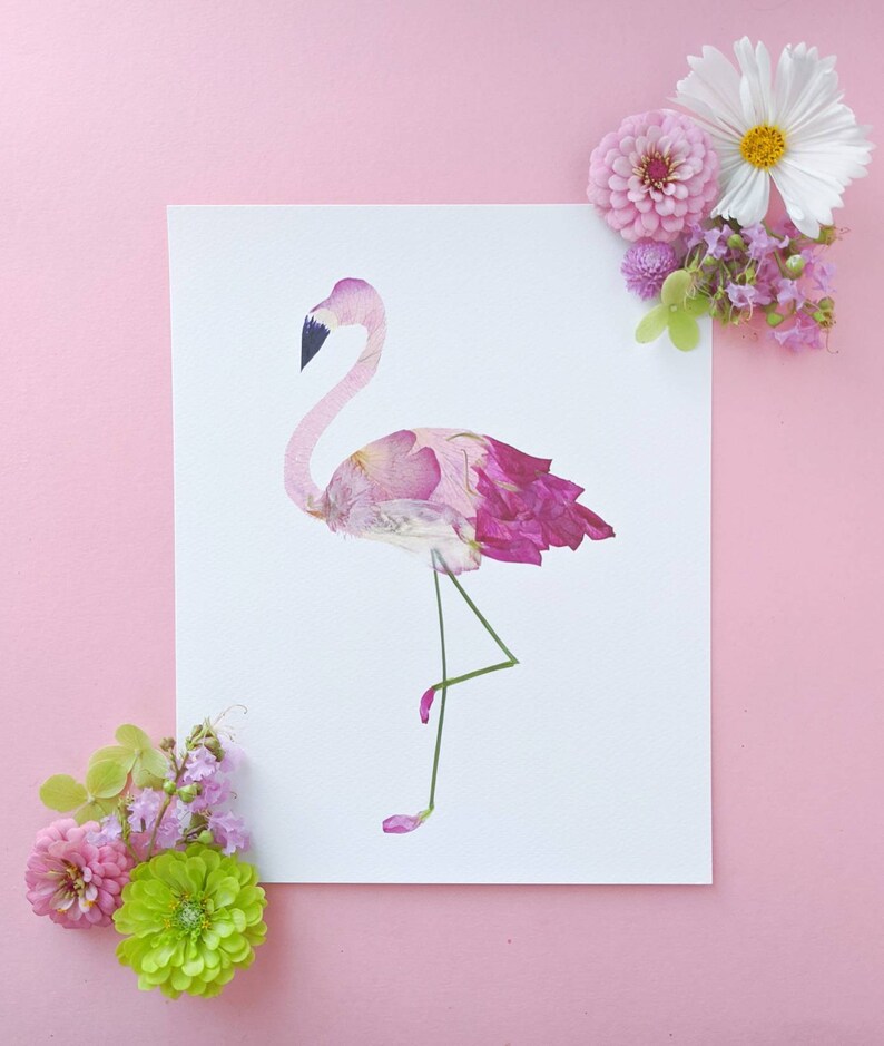 May include: A flamingo art print made from pressed pink and purple flower petals. The flamingo stands on one leg, with a black beak and a pink body. The artwork is on a white background, with pink flowers in the corners.