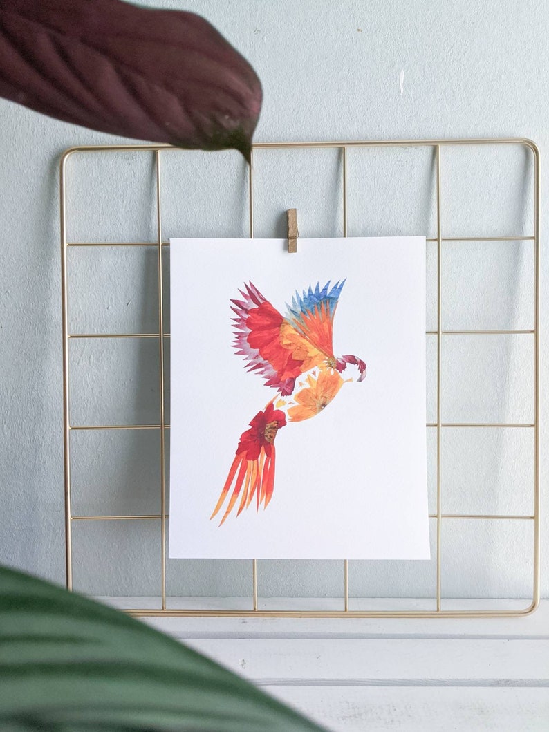 May include: A colorful bird made of pressed flowers, with red, orange, and yellow petals, is flying against a white background. The bird is framed in a gold wire grid.