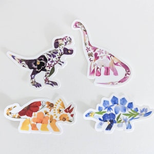 Op de afbeelding: Vier kleurrijke dinosaurus stickers gemaakt van geperste bloemen. De dinosaurussen zijn een Tyrannosaurus Rex, een Stegosaurus, een Triceratops en een Brachiosaurus.