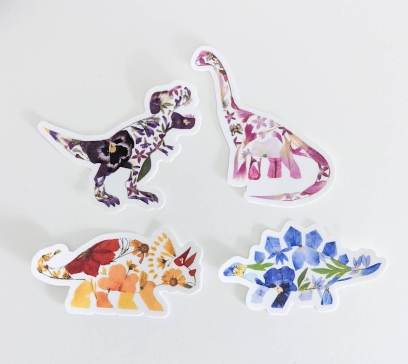 Op de afbeelding: Vier kleurrijke dinosaurus stickers gemaakt van geperste bloemen. De dinosaurussen zijn een Tyrannosaurus Rex, een Stegosaurus, een Triceratops en een Brachiosaurus.