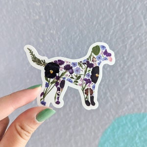 Op de afbeelding: Een witte sticker met een silhouet van een hond gemaakt van geperste bloemen. De bloemen zijn paars, blauw en groen.
