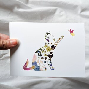 May include: A white card featuring a cat silhouette filled with colorful pressed flowers. The cat is reaching towards a butterfly with pink and yellow wings. The artwork is a unique floral design.