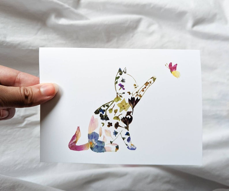 May include: A white card featuring a cat silhouette filled with colorful pressed flowers. The cat is reaching towards a butterfly with pink and yellow wings. The artwork is a unique floral design.