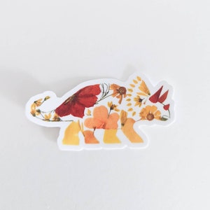Op de afbeelding: Een witte sticker met een bloemmotief van een triceratops. De dinosaurus is gemaakt van geperste bloemen in tinten geel, oranje en rood.