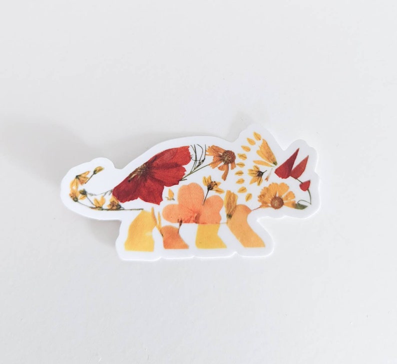 Op de afbeelding: Een witte sticker met een bloemmotief van een triceratops. De dinosaurus is gemaakt van geperste bloemen in tinten geel, oranje en rood.