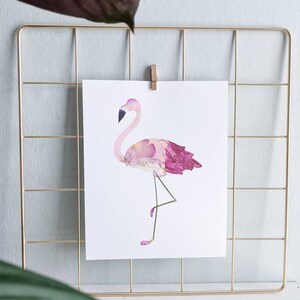 May include: A print of a flamingo made of pink and purple flower petals, displayed on a gold wire grid. The flamingo has a black beak and is standing on one leg. The print is held in place by a wooden clip.