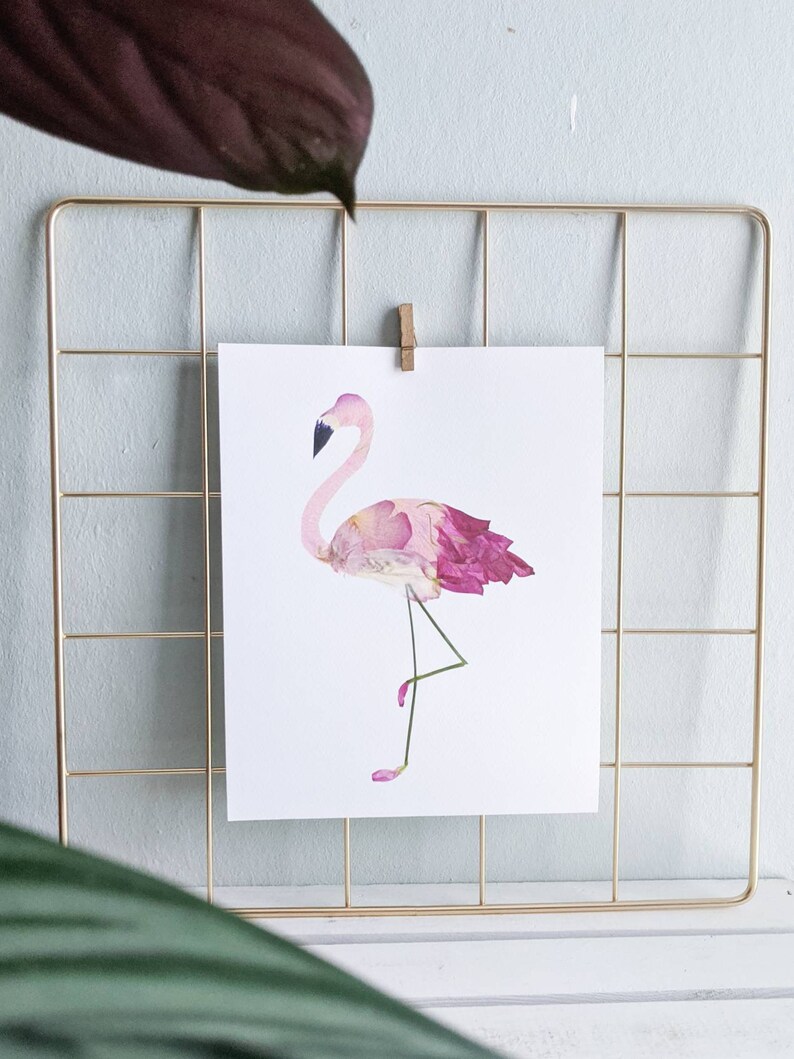 May include: A print of a flamingo made of pink and purple flower petals, displayed on a gold wire grid. The flamingo has a black beak and is standing on one leg. The print is held in place by a wooden clip.