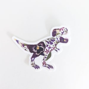 Op de afbeelding: Een paarse en witte bloemen-dinosaurussticker. De dinosaurus is een Tyrannosaurus Rex, en de bloemen zijn geperst en zo gerangschikt dat ze de vorm van de dinosaurus vormen.