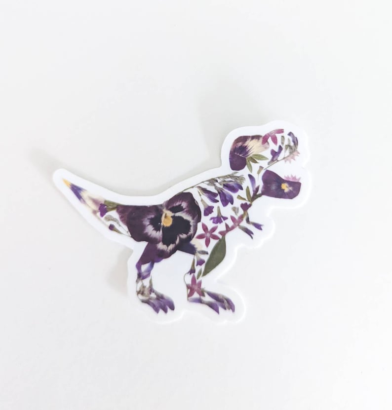 Op de afbeelding: Een paarse en witte bloemen-dinosaurussticker. De dinosaurus is een Tyrannosaurus Rex, en de bloemen zijn geperst en zo gerangschikt dat ze de vorm van de dinosaurus vormen.
