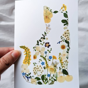 May include: A white card featuring a cat silhouette artfully composed of dried flowers. The floral arrangement includes yellow, white, blue, and purple blooms, creating a delicate and natural aesthetic. The cat shape is clearly defined against the white background.