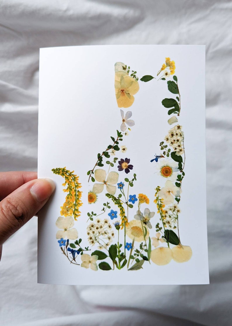 May include: A white card featuring a cat silhouette artfully composed of dried flowers. The floral arrangement includes yellow, white, blue, and purple blooms, creating a delicate and natural aesthetic. The cat shape is clearly defined against the white background.