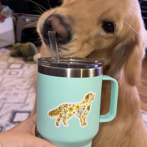Puede incluir: Un vaso Yeti azul claro con tapa y asa de acero inoxidable. El vaso tiene una pegatina de un golden retriever con un patr&oacute;n floral. El vaso est&aacute; siendo sostenido por una mano.