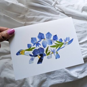 May include: A white card with a blue and yellow pressed flower stegosaurus on it.