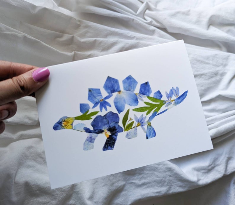 May include: A white card with a blue and yellow pressed flower stegosaurus on it.
