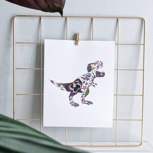 May include: A white print of a Tyrannosaurus Rex made of purple and white flowers. The print is framed in a gold wire grid.