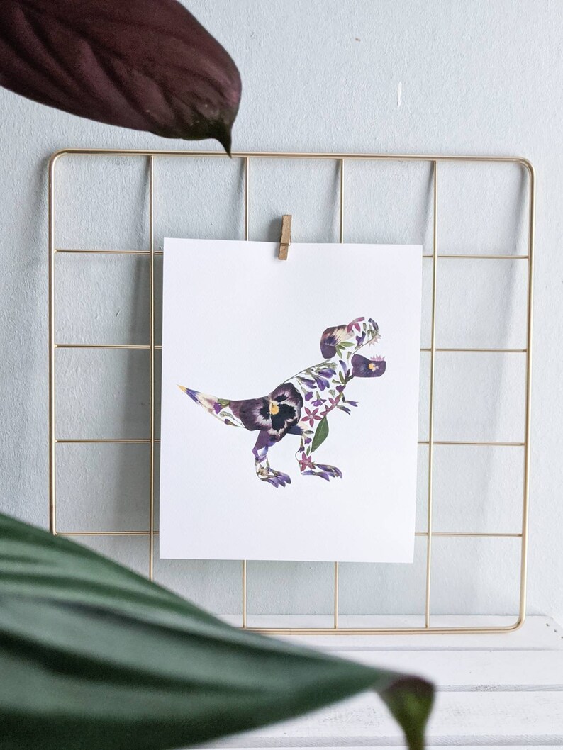 May include: A white print of a Tyrannosaurus Rex made of purple and white flowers. The print is framed in a gold wire grid.