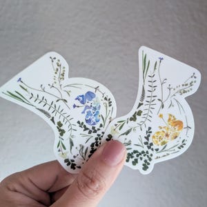 May include: Two white stickers with pressed flowers and leaves. One sticker features blue flowers and the other features yellow flowers.