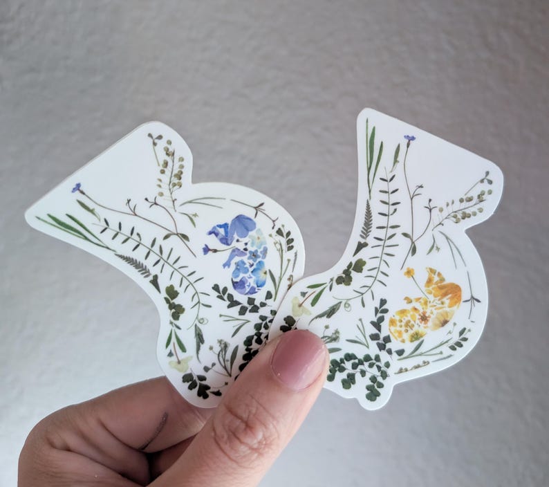May include: Two white stickers with pressed flowers and leaves. One sticker features blue flowers and the other features yellow flowers.