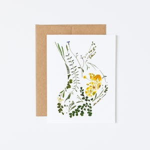May include: A white card with a pressed flower design in shades of green and yellow. The flowers are arranged in the shape of a heart.