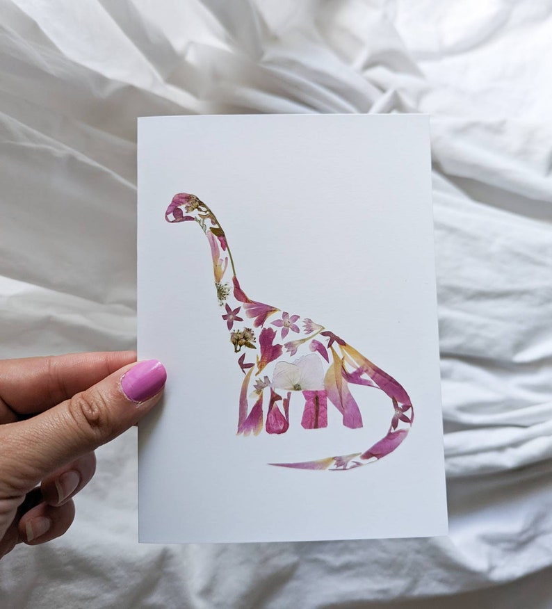 May include: A white card with a pink and white pressed flower dinosaur silhouette.