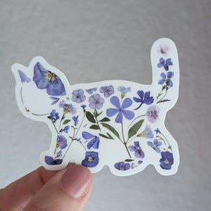 Puede incluir: Una pegatina blanca con una silueta de gato hecha de flores moradas y azules prensadas.