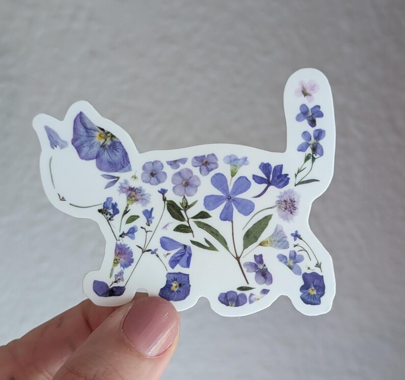 Puede incluir: Una pegatina blanca con una silueta de gato hecha de flores moradas y azules prensadas.