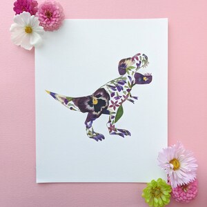 May include: A white card with a T-Rex dinosaur silhouette made from pressed flowers. The dinosaur is purple, pink, and yellow. The card is surrounded by pink and green flowers.