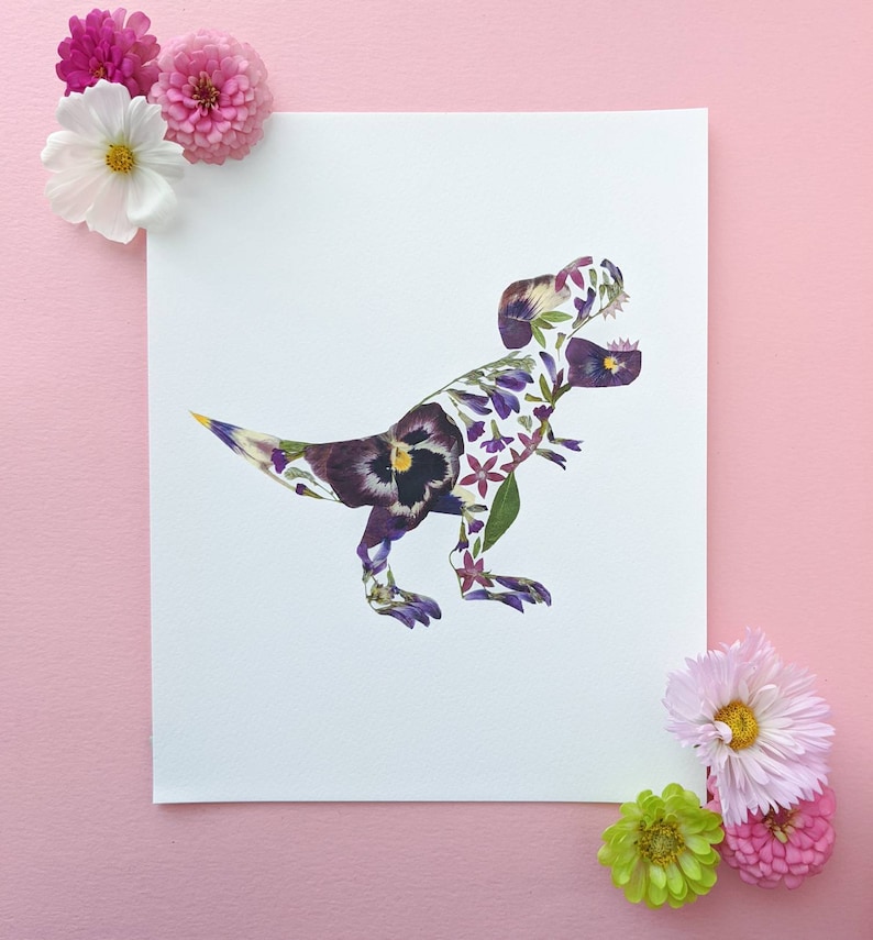 May include: A white card with a T-Rex dinosaur silhouette made from pressed flowers. The dinosaur is purple, pink, and yellow. The card is surrounded by pink and green flowers.