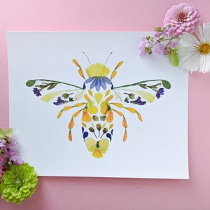 May include: A bee made from pressed flowers. The bee is yellow, blue, and white. The bee is on a white background.