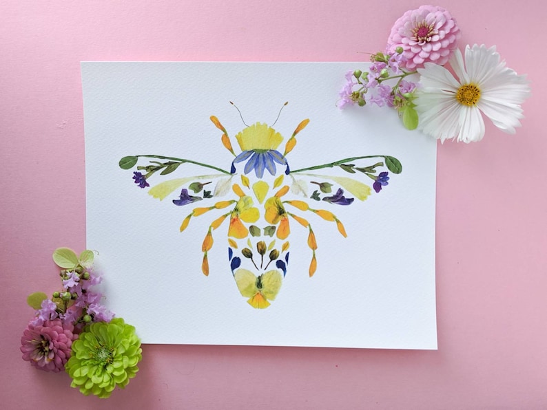 May include: A bee made from pressed flowers. The bee is yellow, blue, and white. The bee is on a white background.